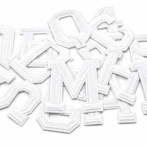 May include: A pile of white embroidered fabric letters, including the letters M, A, S, and Q, on a white background. The letters have a textured look and a stitched border, suggesting they are patches or appliques.