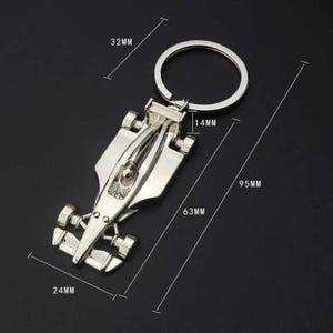 May include: A silver-colored keychain shaped like a race car. The keychain measures 9.5 cm long, 6.3 cm wide, and 3.2 cm tall. The keychain has a ring for attaching keys.