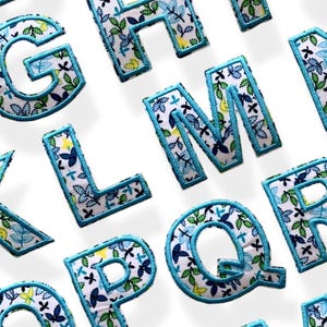 Può includere: Un set di lettere dell'alfabeto in tessuto con un motivo floreale blu e verde. Le lettere sono contornate di blu e hanno uno sfondo bianco.