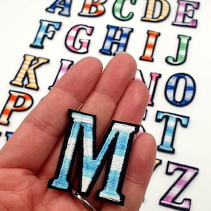 Op de afbeelding: Een set kleurrijke strijkbare alfabetpatches met een gestreept ontwerp. De letter M wordt in het midden van de afbeelding gemarkeerd, met blauwe en witte strepen.