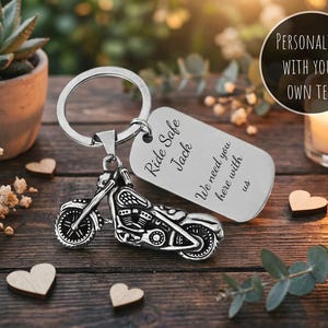 May include: Silver motorcycle keychain with a rectangular tag. The tag is engraved with the words "Ride Safe Jack" and "We need you here with us". The keychain is on a wooden surface with decorative hearts and a succulent plant.