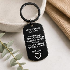 May include: A black metal dog tag keyring featuring white engraved text that reads: LITTLE REMINDERS FOR STACEY, followed by a list of affirmations including You are enough, It's ok to have bad days, Dont believe everything you think, You are so very loved, Be kinder to yourself, and LOVE MUM x with a heart icon.