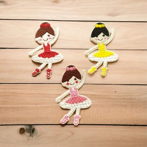 May include: Three embroidered iron-on patches of ballerinas. The ballerinas are wearing pink, red, and yellow tutus. Each ballerina has brown hair and a white background.
