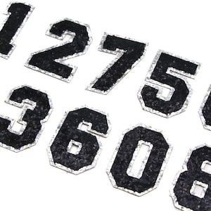 May include: A set of black iron-on numbers with a silver glitter outline. The numbers are 1, 2, 7, 5, 3, 6, 0, and 8.