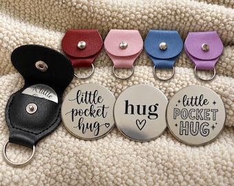 Puede incluir: Una colección de pequeñas fichas metálicas circulares con texto grabado en negro, incluyendo "little pocket hug" y "hug" con símbolos de corazón, junto a fundas de cuero a juego en negro, rojo, rosa, azul y morado, con un botón de presión.