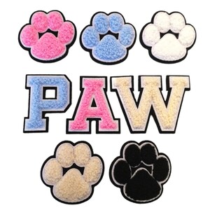 May include: A set of six iron-on patches featuring paw prints and the word "PAW" in various colours. The paw prints are in pink, blue, white, beige, and black. The letters are in blue, pink, and beige.