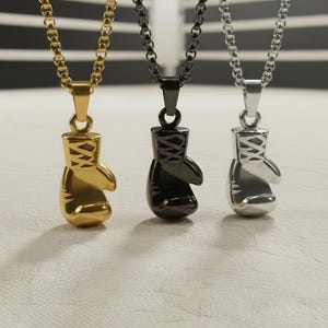 Puede incluir: Tres collares con colgantes de guantes de boxeo en oro, negro y plata. Cada colgante es un guante de boxeo en miniatura detallado, suspendido de una cadena a juego. El fondo es una superficie texturizada neutra.