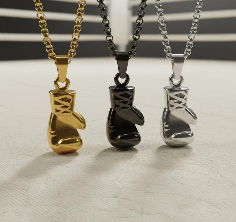 Puede incluir: Tres collares con colgantes de guantes de boxeo en oro, negro y plata. Cada colgante es un guante de boxeo en miniatura detallado, suspendido de una cadena a juego. El fondo es una superficie texturizada neutra.