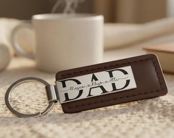 May include: A rectangular brown leather keychain featuring a silver-toned metal plate engraved with the word DAD in large black letters. The names Megan, Rhys, and Etta are inscribed in a cursive script across the center of the letters.
