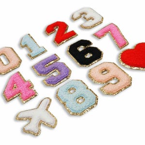 Puede incluir: Un conjunto de diez parches de fieltro en varios colores, cada uno con un número diferente del 0 al 9. Los parches están delineados con purpurina dorada y tienen una textura peluda. También hay un parche de corazón de fieltro rojo con purpurina dorada que lo delinea.