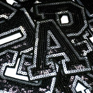 Peut inclure: Gros plan sur des lettres en tissu noir et argent&eacute; &agrave; paillettes. Les lettres sont dispos&eacute;es de mani&egrave;re al&eacute;atoire et cousues ensemble. Le tissu est brillant et r&eacute;fl&eacute;chissant.