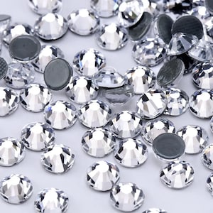 May include: A large quantity of clear glass rhinestones, some with adhesive backing, scattered on a white surface.