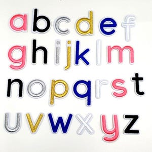 Puede incluir: Un conjunto de 26 letras del alfabeto termoadhesivas de colores. Las letras son de varios colores, incluyendo rosa, negro, dorado, plateado, azul y blanco. Las letras son todas en mayúsculas y están ordenadas alfabéticamente.