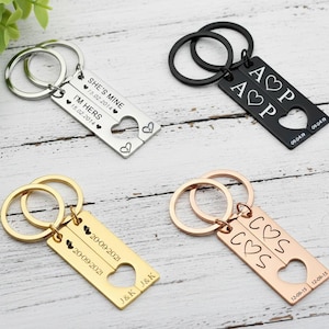 May include: Four keychains with rectangular pendants and heart cutouts. One is silver with the text "SHE'S MINE" and "I'M HERS" with dates. Another is black with initials "A P" and a date. The third is gold with initials "J & K" and a date. The fourth is rose gold with initials "C S" and a date.