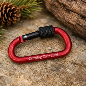 May include: A red aluminum carabiner with a black screw-gate locking mechanism. The metal frame features the white engraved text "Camping Tour 2026" along the bottom curve. The carabiner has a smooth, matte finish and a functional spring-loaded gate.