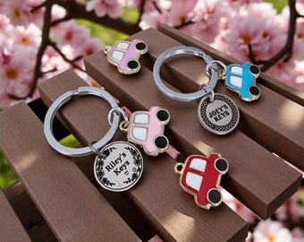 May include: Two silver metal keychains featuring enamel car charms in pink, blue, and red. One keychain has a round tag engraved with 'Riley's Keys' and a floral border, while the other has a round tag engraved with 'JOEY'S KEYS' and a dotted border.