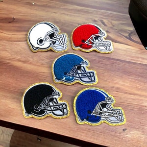 Op de afbeelding: Vijf geborduurde patches van American footballhelmen in wit, rood, blauw, zwart en donkerblauw. Elke patch heeft een gouden rand en gedetailleerde stiksels, die het gezichtsmasker en het ontwerp van de helm laten zien. Ideaal voor sportliefhebbers.