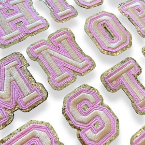 May include: A set of pink and gold glitter iron-on letters. The letters are in a varsity style font and are perfect for adding a touch of sparkle to any project.