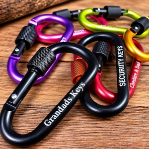 May include: A collection of colorful metal carabiners with screw-lock gates. The black carabiners feature white text reading Grandads Keys and SECURITY KEYS, while a red one is engraved with Cheshire & Dad. The set includes purple, lime green, and gold.