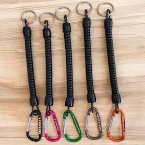 Könnte beinhalten: Fünf schwarze, einziehbare Schlüsselanhänger mit silbernen Schlüsselringen und farbigen Karabinerhaken. Die Haken sind schwarz, rosa, grün, grau und orange. Jeder Haken hat einen anderen Namen oder Text: "SECURITY", "Lily-anne", "Keep Adventuring!", "Jake's Keys" und "ICE: 07000XXXXXX".