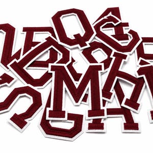 May include: A collection of maroon varsity-style letter and number patches, each featuring a bold, block-style font with a thick white border. The embroidered patches are arranged in a scattered pile, showcasing various characters including M, Q, and 8.