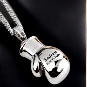 Puede incluir: Colgante de guante de boxeo plateado con una cadena. El colgante tiene el nombre "Andrew" y la frase "Born To Fight" grabados. La cadena es de tipo barbada plateada. El fondo es negro.
