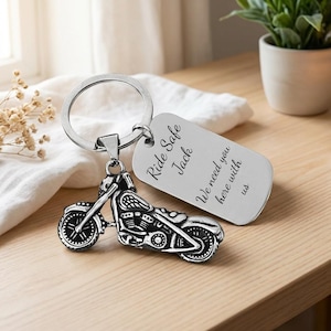 May include: A silver motorcycle keychain with a detailed bike charm and a rectangular tag. The tag is engraved with the words "Ride Safe Jack, We need you here with us". The keychain is on a wooden surface.