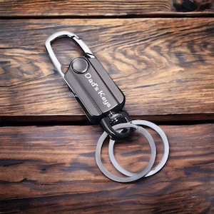 May include: A black metal keychain with a silver carabiner and two silver rings. The keychain is engraved with the words "Dad's Keys".