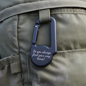 Op de afbeelding: Een matzwarte metalen karabijnhaak met een veerbelaste sluiting en een afgeronde basis. Het oppervlak is gegraveerd met de witte cursieve tekst, So you always find your way home!, bevestigd aan een lus op een olijfgroene stoffen ondergrond.