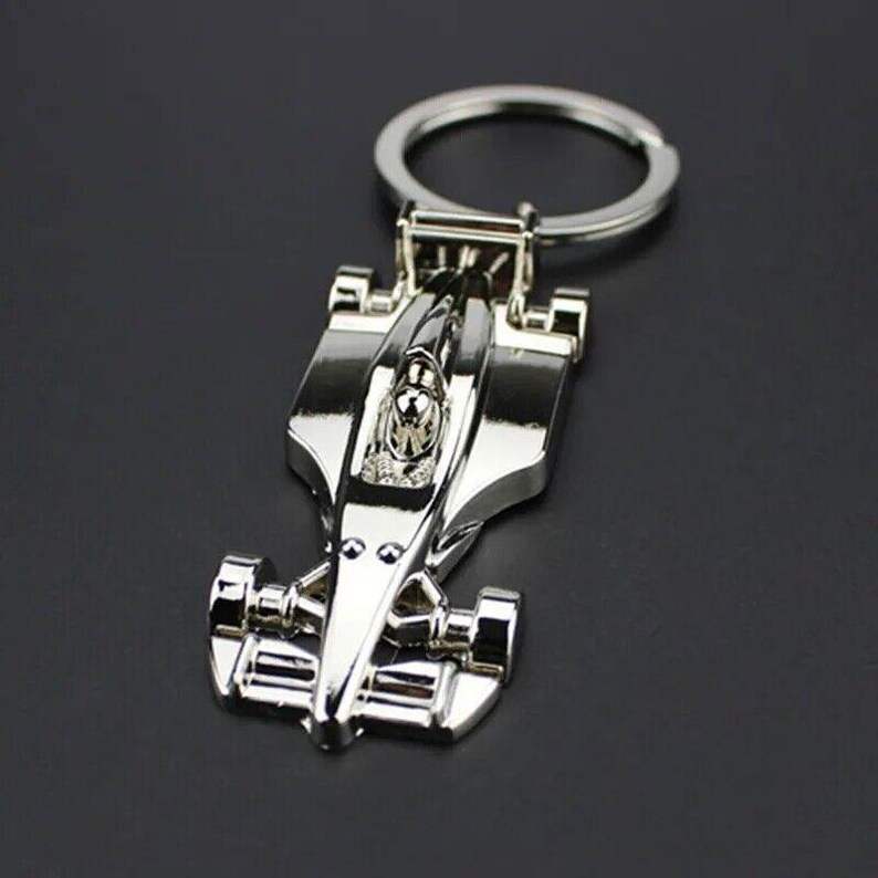 May include: A silver metal keychain featuring a detailed model of a race car. The car has a driver in the cockpit and is attached to a silver key ring.
