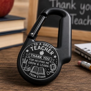 Op de afbeelding: Een zwarte karabijnhaak met een rond middenstuk voorzien van een witte gravure met de tekst TO A GREAT TEACHER THANK YOU FOR HELPING ME GROW & LEARN FROM ETHAN, vergezeld door iconen van een appel, potloden, boeken, een wereldbol en een hart.