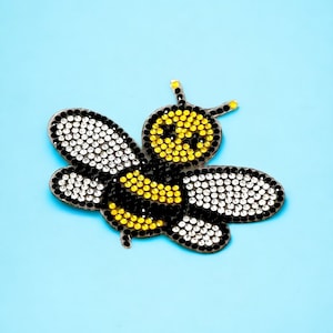 Puede incluir: Una abeja amarilla y negra con detalles de pedrería transparente. La abeja está hecha de tela y tiene un efecto 3D.