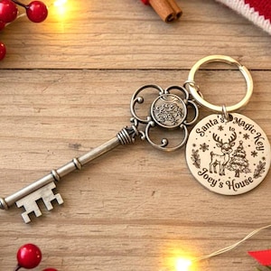 Könnte beinhalten: Antiker silberfarbener Schlüssel mit einem runden Anhänger mit der Aufschrift "Santa's Magic Key" mit einem Rentier- und Weihnachtsbaum-Design. Auf dem Anhänger steht auch "Joey's House". Der Schlüssel liegt auf einer Holzoberfläche.