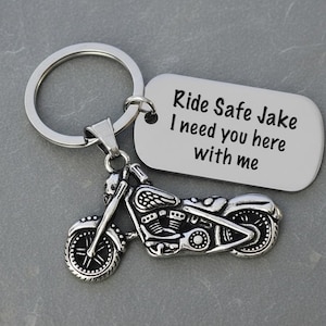 May include: A silver motorcycle keychain with a matching tag. The tag reads "Ride Safe Jake I need you here with me". The motorcycle charm has detailed features, including wheels, a seat, and handlebars. The keychain is on a gray surface.
