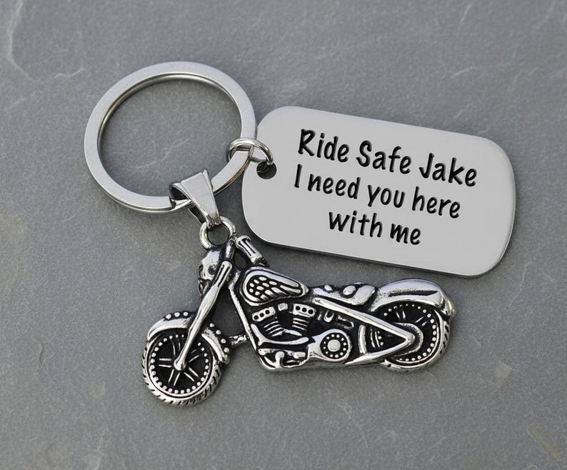 May include: A silver motorcycle keychain with a matching tag. The tag reads "Ride Safe Jake I need you here with me". The motorcycle charm has detailed features, including wheels, a seat, and handlebars. The keychain is on a gray surface.
