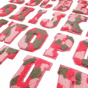 May include: A set of pink and green camouflage felt letters. The letters are in a variety of sizes and are arranged in a random order.