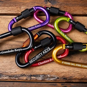 May include: A collection of colourful metal carabiners with screw-gate locking mechanisms. The carabiners are engraved with various text including Studio Keys, Jo's Keys, Grandads Keys, SECURITY KEYS, Cheshire & I, and IF FOUND CALL 07890 123456.