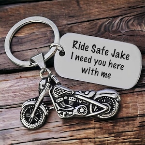 May include: Silver motorcycle keychain with a rectangular tag. The tag reads "Ride Safe Jake I need you here with me". The motorcycle charm is detailed with wheels, engine, and handlebars. The keychain is on a wooden surface.