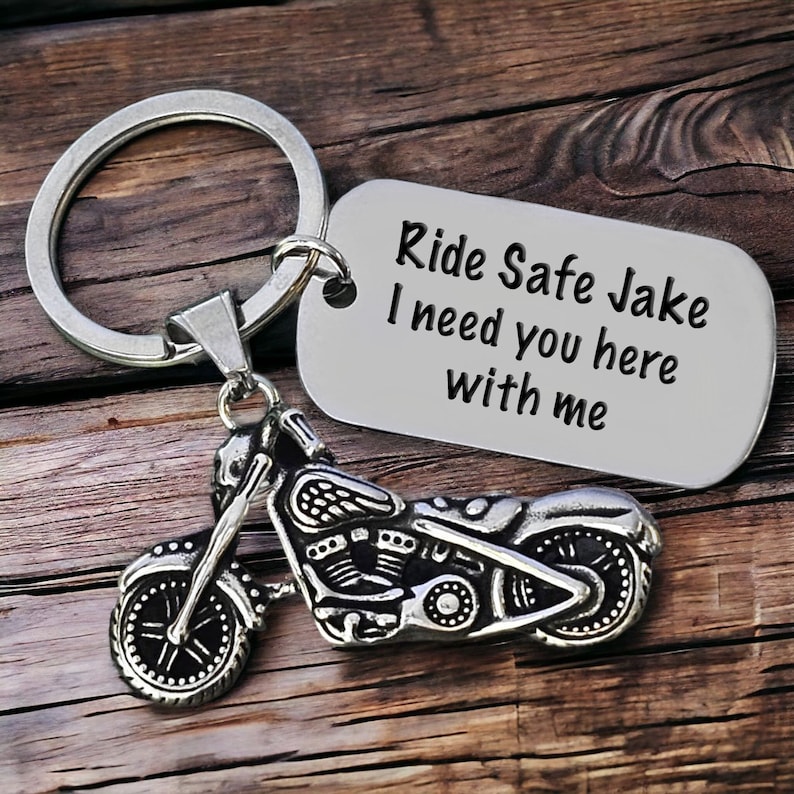 May include: Silver motorcycle keychain with a rectangular tag. The tag reads "Ride Safe Jake I need you here with me". The motorcycle charm is detailed with wheels, engine, and handlebars. The keychain is on a wooden surface.