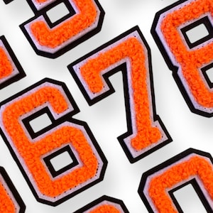 Op de afbeelding: Een set oranje en zwarte viltcijfers, waaronder de cijfers 1, 2, 3, 4, 5, 6, 7, 8 en 0. De cijfers zijn zwart omrand en hebben een witte rand.