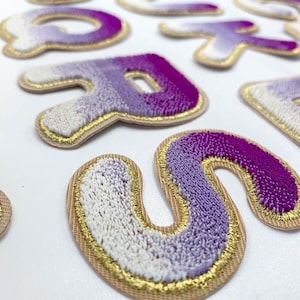May include: A set of colourful felt letters with a gradient of purple, white and pink. The letters are outlined in gold thread and have a fuzzy texture.