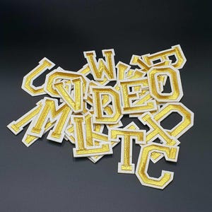 May include: A pile of gold-coloured felt letters with white outlines. The letters are in various sizes and are arranged in a random order. The letters are all uppercase and include the letters C, V, I, D, E, O, M, L, T, and W.