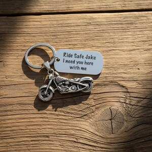 May include: A silver motorcycle keychain with a tag that reads "Ride Safe Jake I need you here with me." The keychain is on a wooden surface with visible grain and shadows.