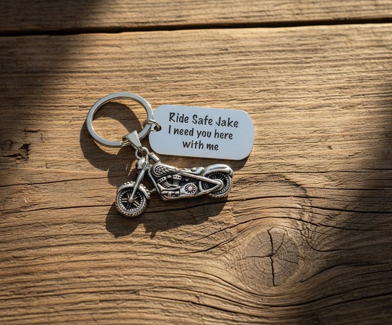 May include: A silver motorcycle keychain with a tag that reads "Ride Safe Jake I need you here with me." The keychain is on a wooden surface with visible grain and shadows.