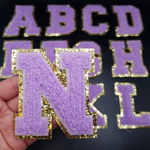 May include: Purple felt letter patches with gold glitter trim. The letters are in a block font style. The letters shown are A, B, C, D, I, H, N, K, and L.