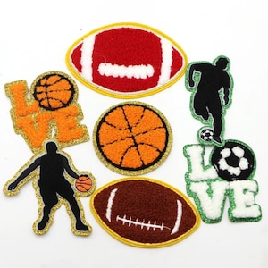 May include: Eight iron-on patches featuring different sports themes. The patches are made of fuzzy fabric and have a gold glitter outline. The patches include a football, basketball, soccer ball, and a silhouette of a basketball player and a soccer player. Two patches say "LOVE" in white letters with a fuzzy green background.
