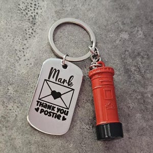Könnte beinhalten: Ein silberner Schlüsselanhänger mit einem Anhänger mit dem Namen "Mark", einer Umschlag-Grafik mit einem Herz und den Worten "THANK YOU POSTIE". Ein roter Miniatur-Postkasten-Anhänger ist befestigt. Der Schlüsselanhänger befindet sich an einem silbernen Ring.