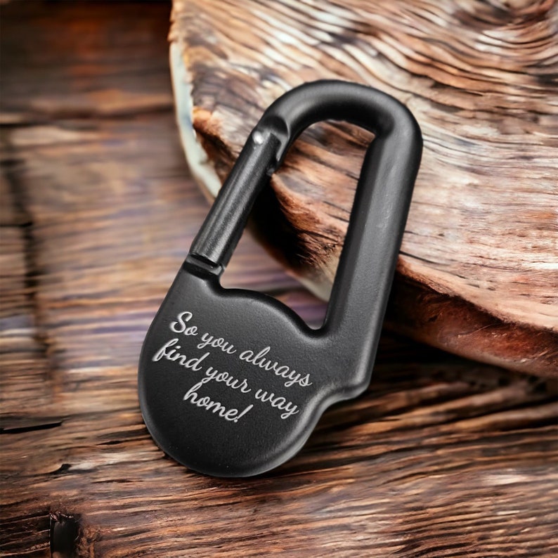 Op de afbeelding: Een matzwarte karabijnhaak sleutelhanger met een veerbelaste sluiting en een afgeronde basis. Het oppervlak is gegraveerd met de witte tekst "So you always find your way home!" in een cursief lettertype, wat zorgt voor een duidelijke boodschap.