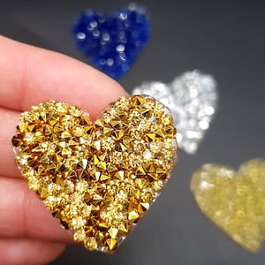 May include: A gold heart-shaped embellishment with a sparkly, rhinestone design. The heart is made of clear resin and is decorated with small, gold-colored rhinestones.