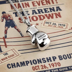 Puede incluir: Colgante de guante de boxeo plateado en una cadena. El guante est&aacute; grabado con "Andrew Born To Fight". El fondo presenta gr&aacute;ficos tem&aacute;ticos de boxeo, incluyendo el texto "MAIN EVENT" y "CHAMPIONSHIP BOUT OCT 26, 1970."